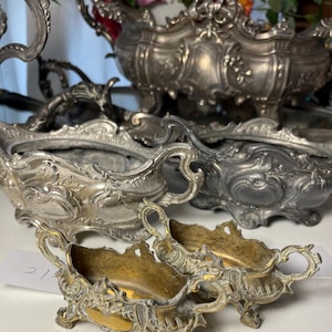 Puede incluir: Un surtido de fuentes y cuencos decorativos de color plateado antiguo. Las piezas presentan detalles ornamentados, incluyendo asas enrolladas y motivos florales. Algunos tienen un interior dorado. La colección se exhibe sobre una superficie blanca.