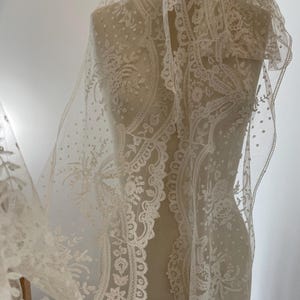 May include: An off-white lace veil featuring intricate floral embroidery and a delicate dotted pattern throughout the sheer fabric. The lace has a scalloped edge with detailed botanical motifs, draped over a dress form mannequin.