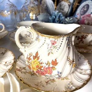 Puede incluir: Un juego de jarrita y platillo de porcelana blanca con un diseño floral en amarillo, rosa y naranja. La jarrita tiene un borde y asa dorados, y el platillo tiene un borde festoneado. Parte de un juego de té vintage.