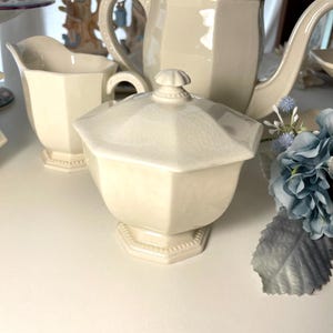 Pode incluir: Um açucareiro de cerâmica na cor creme com tampa a condizer, apresentando uma forma octogonal facetada e um acabamento de esmalte craquelado subtil. O açucareiro assenta numa base de pedestal e inclui um pequeno puxador decorativo na tampa.