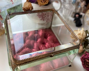 May include: A vintage glass jewelry box with a red velvet interior. The box has a clear glass top and sides, with a brass-colored metal frame. The interior features a tufted velvet cushion. The box is square-shaped and sits on small feet.