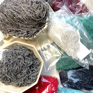 Peut inclure: Un assortiment de brins de perles de différentes couleurs, dont argent, rouge, blanc, sarcelle et noir. Les perles sont emballées dans des sacs en plastique transparents et présentées dans des plats en céramique blancs.