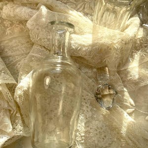 May include: Clear glass bottles of various shapes and sizes rest on a cream-colored lace fabric. The bottles include a tall, cylindrical bottle, a small, diamond-shaped bottle, and a rectangular bottle with a stopper. The scene is bathed in soft light.