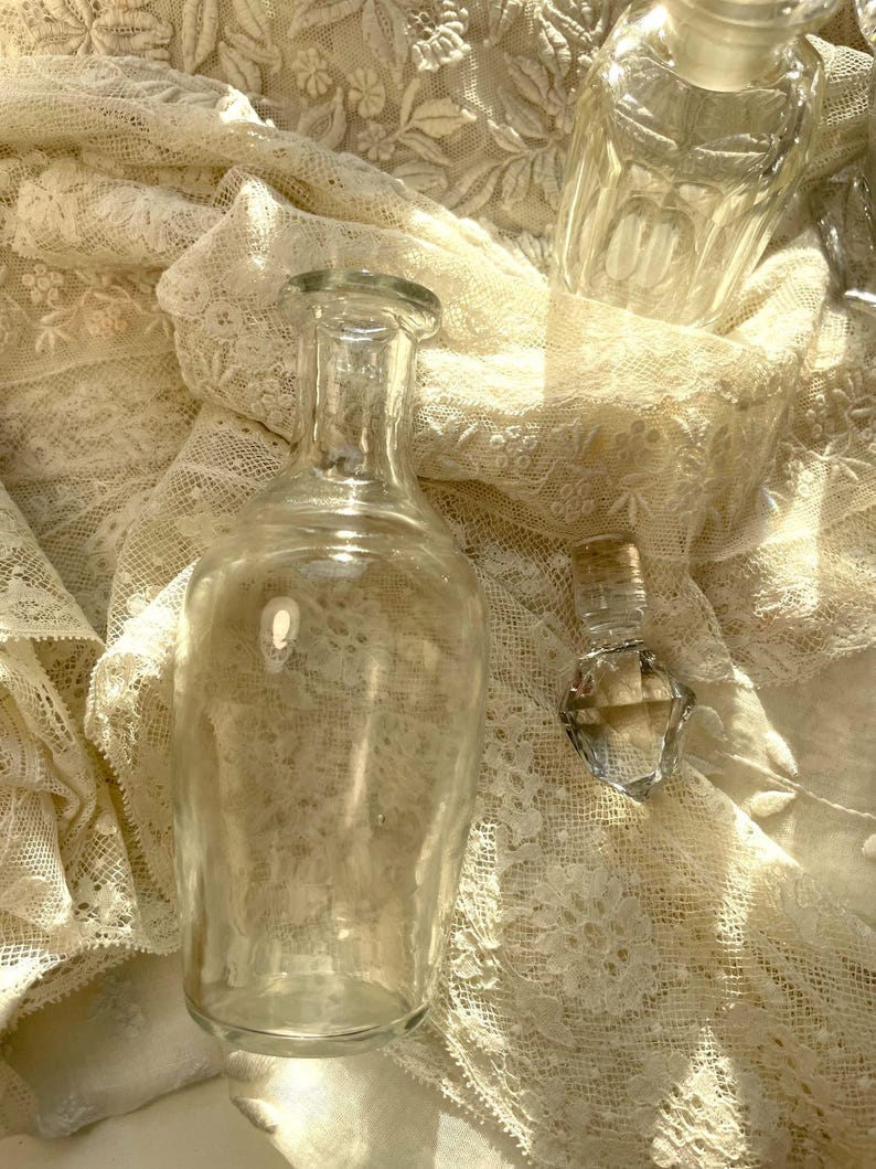 May include: Clear glass bottles of various shapes and sizes rest on a cream-colored lace fabric. The bottles include a tall, cylindrical bottle, a small, diamond-shaped bottle, and a rectangular bottle with a stopper. The scene is bathed in soft light.