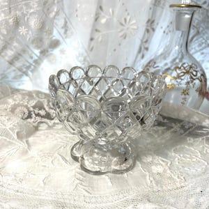May include: A clear, cut-glass bowl with a scalloped rim and diamond pattern. The bowl sits on a pedestal base. A decorative glass bottle with gold accents is in the background. The items are on a white lace surface.