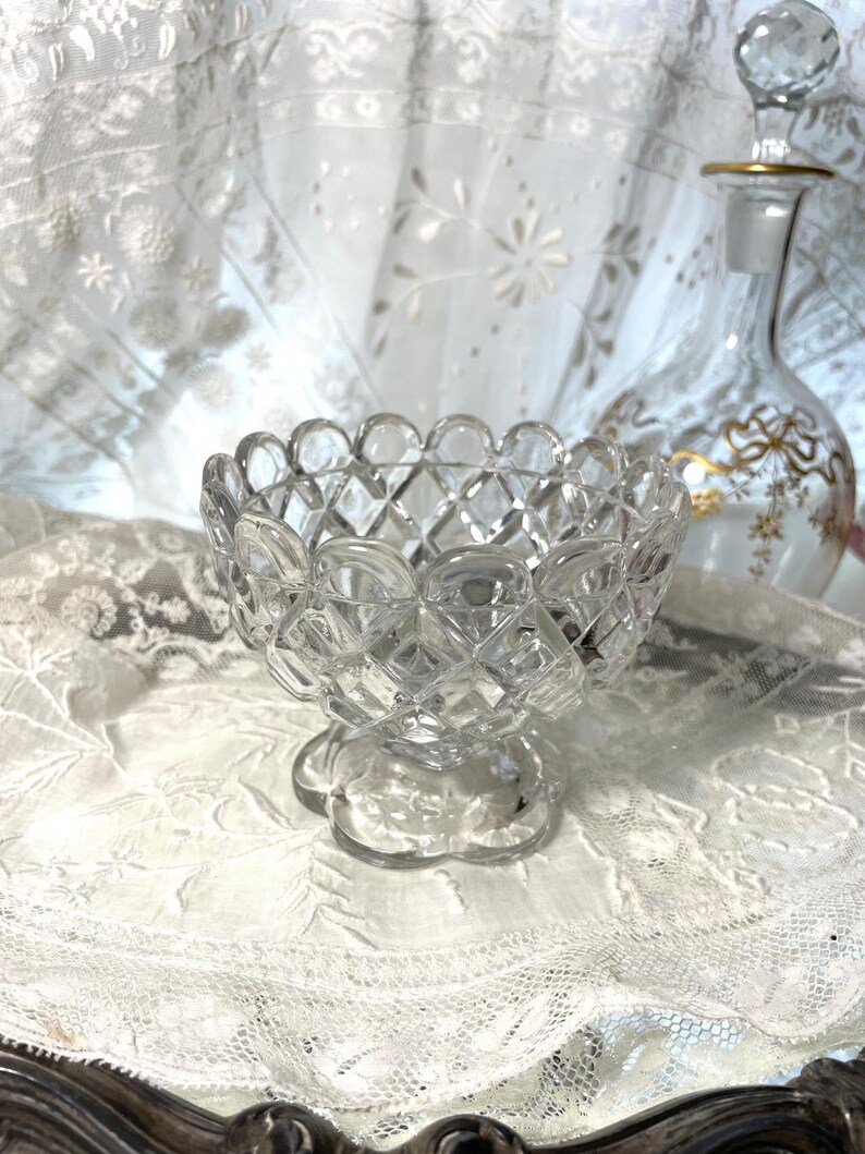 May include: A clear, cut-glass bowl with a scalloped rim and diamond pattern. The bowl sits on a pedestal base. A decorative glass bottle with gold accents is in the background. The items are on a white lace surface.