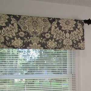 May include: A valance with a black and white floral pattern hangs above a window with white blinds. The valance is made of a soft, textured fabric.