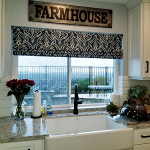 May include: A white kitchen sink with a black tap, a white worktop, and a window with a blue and white patterned roman blind. The window has a view of a garden with a swimming pool and a patio. The sign above the window reads "FARMHOUSE".