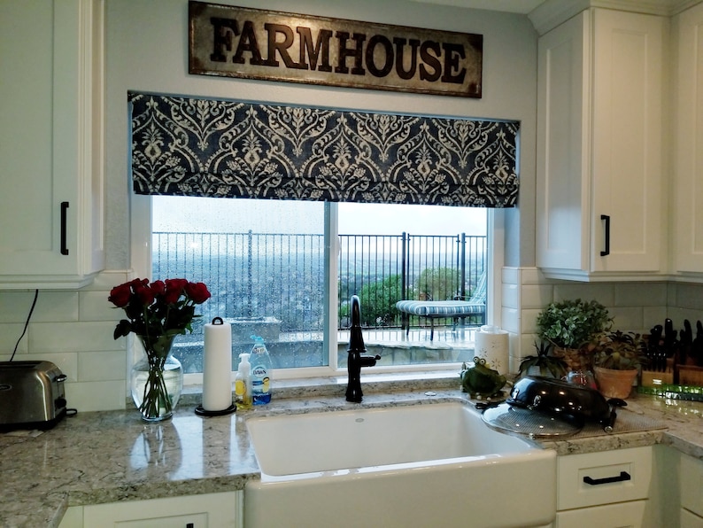 May include: A white kitchen sink with a black tap, a white worktop, and a window with a blue and white patterned roman blind. The window has a view of a garden with a swimming pool and a patio. The sign above the window reads "FARMHOUSE".