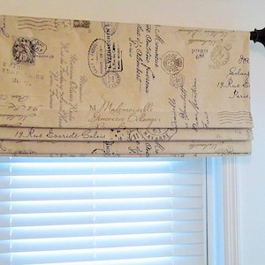 May include: Beige Roman shade with a vintage postcard design featuring stamps, script, and French text. The shade is mounted above a white window with horizontal blinds. The rod is dark brown.