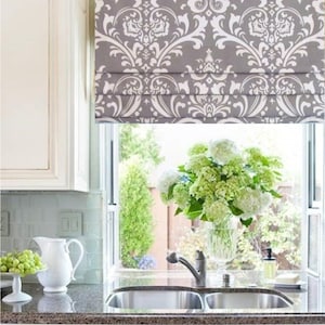 May include: A gray and white patterned roman shade hangs above a kitchen sink with a double basin. A vase of white flowers sits on the windowsill.