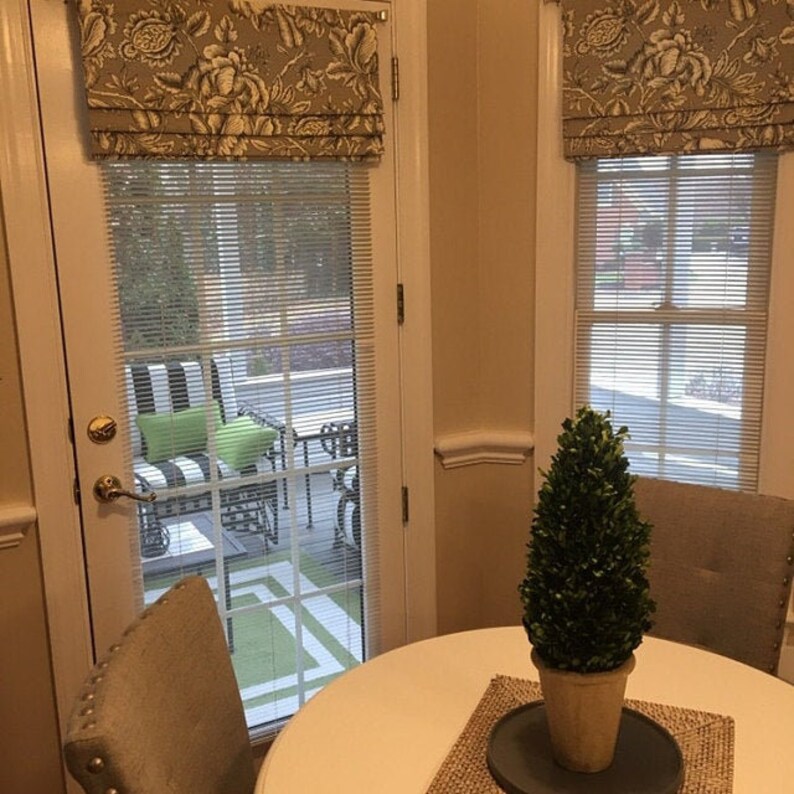 May include: A white table with a green plant in a brown pot on top of a woven placemat. The table is in front of a window with white blinds and a floral valance. There is a white chair to the left of the table.