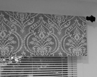 May include: A gray and white patterned valance with a decorative finial on the right side. The valance is hanging above a window with white blinds.