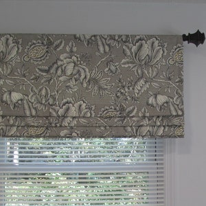 May include: A gray and white floral patterned roman shade with a black metal rod. The shade is mounted above a white window with white blinds.