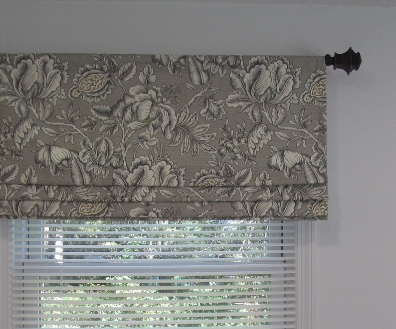 May include: A gray and white floral patterned roman shade with a black metal rod. The shade is mounted above a white window with white blinds.