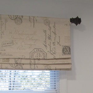 May include: A beige roman shade with a vintage French postage stamp print. The shade is pulled up and the fabric is gathered in pleats. The shade is mounted on a white wall with a brown metal finial.