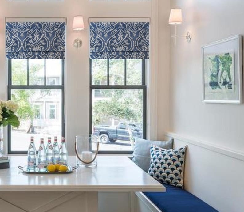 May include: A white kitchen table with a blue bench seat. The table has a glass vase with lemons and a tray of water bottles. The windows have blue and white patterned roman blinds.