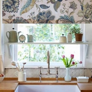 May include: A white kitchen window with a floral valance, a white shelf with a teapot, a clock, and a pitcher, a white sink with a chrome faucet, and a vase of pink tulips.