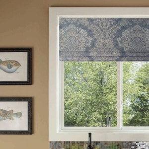 May include: A kitchen window with a white frame and a blue patterned roman shade. The window is open and shows a view of green trees outside. There is a yellow pot on the counter below the window.