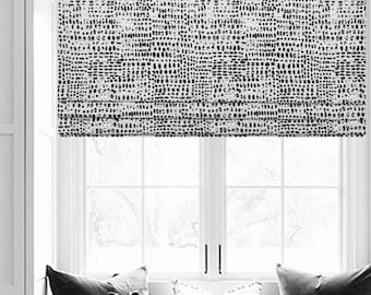 May include: A black and white patterned roman shade with a white background and black abstract shapes. The shade is raised and the window behind it is visible.