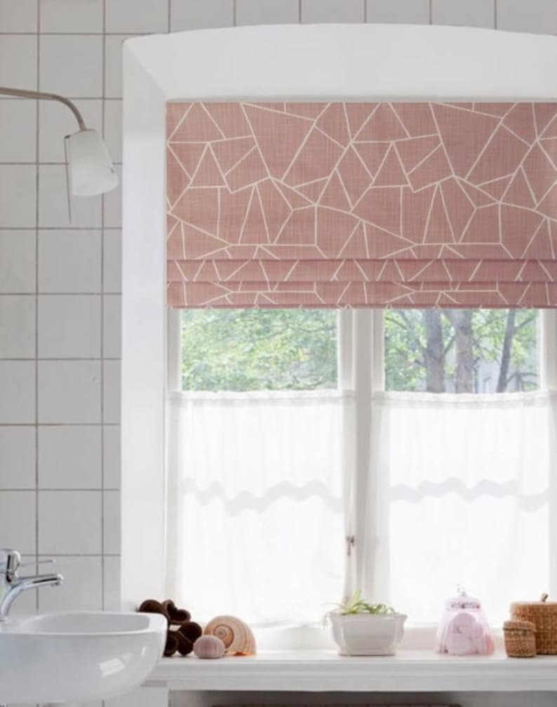 Op de afbeelding: Een roze rolgordijn met een geometrisch patroon van witte lijnen. Het rolgordijn is gedeeltelijk omhooggetrokken, waardoor een wit transparant gordijn met gegolfde randen zichtbaar wordt. Het raam bevindt zich in een badkamer met witte betegelde muren en een witte wastafel.
