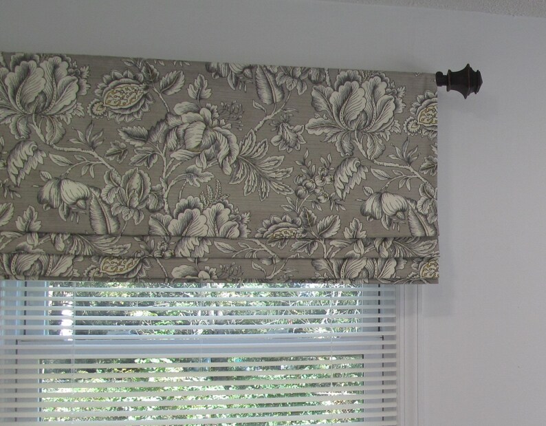 May include: A gray and white floral patterned roman shade with a brown metal rod. The shade is pulled up to reveal a white window blind.