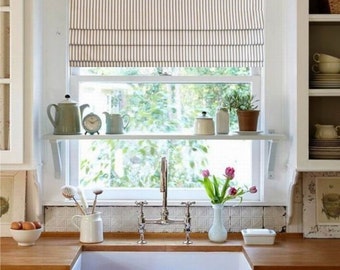 Puede incluir: Escena de cocina luminosa con un fregadero blanco de granja, encimeras de madera y una ventana con una persiana romana a rayas. Una estantería exhibe teteras, un reloj y plantas. Un jarrón de tulipanes rosas añade color.