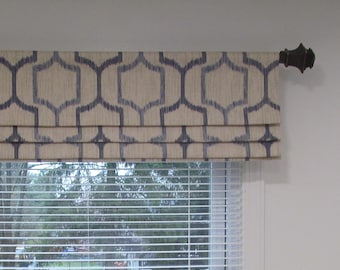 May include: A beige and blue patterned roman shade with a dark brown metal rod. The shade is pulled up to reveal a white window blind.