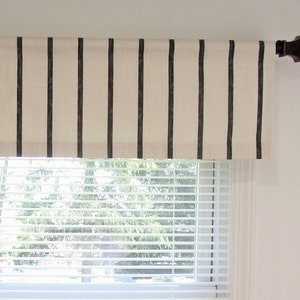 May include: A window valance with a cream-colored background and vertical black stripes. The valance is mounted on a dark brown decorative rod. The window below has white blinds.