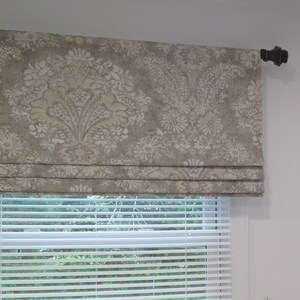 May include: A beige roman shade with a white floral pattern hangs on a white wall above a white window blind.