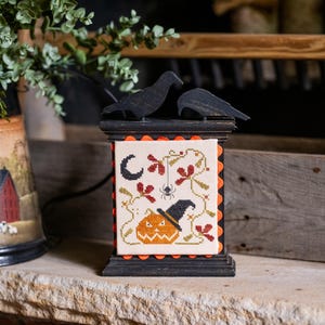 May include: A rectangular black wooden frame featuring a cross-stitch panel with a pumpkin wearing a witch hat, a spider, and a crescent moon. Two black bird silhouettes sit atop the frame, which is accented with orange scalloped trim.
