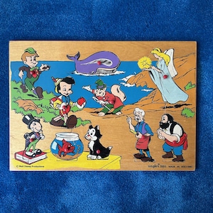 May include: Wooden jigsaw puzzle featuring Disney characters, including Pinocchio, a whale, and a cat. The puzzle has a light brown background with colorful illustrations of various characters and scenes. The text "Walt Disney Productions" and "simplex toys MADE IN HOLLAND" are visible.