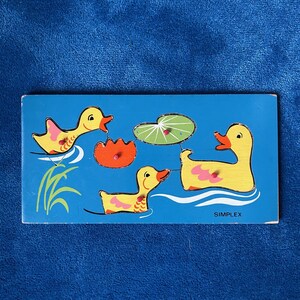 Può includere: Un puzzle in legno vintage con tre anatre gialle che nuotano in uno stagno blu, con una ninfea e un fiore arancione. Il puzzle è etichettato "SIMPLEX" e presenta un design semplice e colorato.