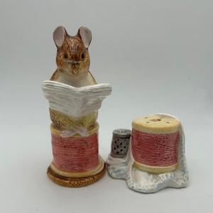 May include: A ceramic salt and pepper shaker set featuring a mouse reading a newspaper atop a spool of thread. The set includes a thimble and a second spool of thread, both with red thread and cream-colored accents.