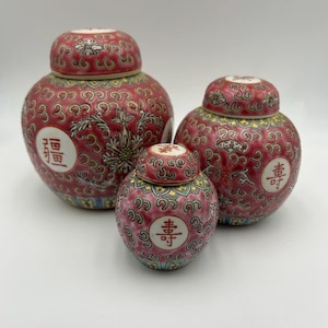 Peut inclure: Trois pots à gingembre ronds en céramique avec couvercles, présentant un fond rose foncé avec des motifs complexes de volutes dorées, vertes et bleues. Chaque pot affiche un médaillon circulaire blanc avec des caractères chinois rouges.