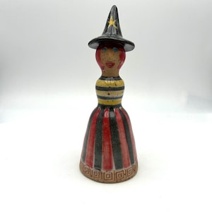 May include: A ceramic figurine of a figure wearing a black pointed hat with a yellow star, red hair, and blue eyes. The torso features yellow and black horizontal stripes, while the skirt has vertical red and black stripes with a geometric border.