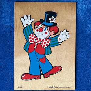May include: Wooden puzzle featuring a colorful clown with a red nose, bow tie, and hat. The clown is wearing blue pants, a red vest, and a green and blue striped jacket. The puzzle is made by Simplex Toys in Holland. The puzzle is on a wooden board.