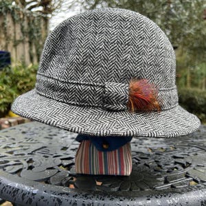 Könnte beinhalten: Ein grau-weißes Cloche-Hut mit Fischgrätenmuster und kleinem Rand. Der Hut hat eine dekorative Schleife und eine Ansammlung brauner und roter Federn. Der Hut wird auf einem dekorativen Ständer präsentiert.