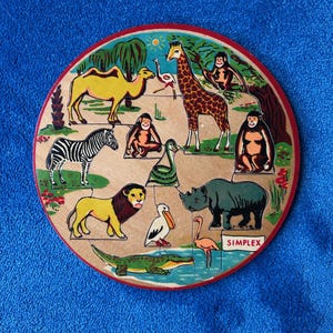 May include: A wooden animal puzzle featuring a camel, zebra, lion, giraffe, rhino, crocodile, and other animals. The puzzle is round with a red border and the word "SIMPLEX" is printed on the bottom right.