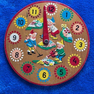 May include: A round wooden clock face featuring a red rotating hand and numbers one through twelve, each surrounded by a colorful floral border. The center displays three illustrated gnomes gardening with a rake, a watering can, and flowers.