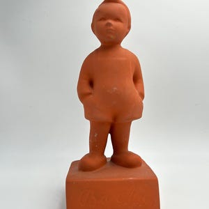 May include: A terracotta-colored figurine of a young child with hands in pockets, standing on a rectangular base. The figure has a simple, stylized design with a small, upright hairstyle. The base has an inscription.
