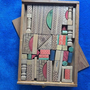 Könnte beinhalten: Eine rechteckige Holzkiste gefüllt mit verschiedenen Holzbausteinen. Die Bausteine weisen aufgedruckte Muster auf, darunter Ziegeltexturen, Bögen und geometrische Designs in Rot, Grün, Blau und natürlichen Holztönen. Ein Baustein trägt den Namen Peter.