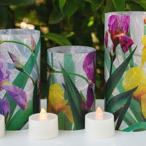 May include: Three white pillar candles with a floral design featuring purple and yellow irises and green leaves. The candles are lit with tea lights.
