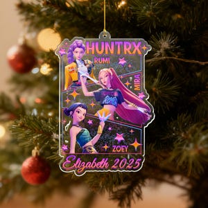 May include: A clear acrylic Christmas ornament featuring three animated female characters. The ornament has the text "HUNTRX" at the top, with the names Rumi, Mira, and Zoey. The name "Elizabeth 2025" is written in cursive at the bottom. The ornament is hanging on a Christmas tree.