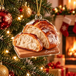 May include: A clear acrylic Christmas tree ornament featuring a high-resolution print of a sliced loaf of sourdough bread on a wooden cutting board. The name Emma is printed in white script on the board. It hangs from a gold cord loop.