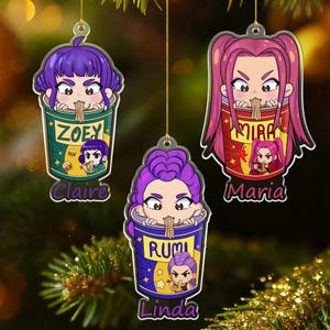 Peut inclure: Trois décorations de Noël de style anime. Chaque décoration représente un personnage regardant par-dessus le bord d'un bol de ramen. Les bols sont étiquetés avec des noms comme Zoey, Mira et Rumi. Les décorations sont suspendues à des cordes dorées.