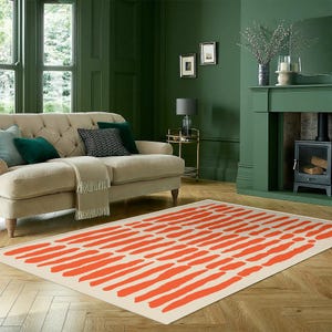 Könnte beinhalten: Rechteckiger Teppich mit cremefarbenem Hintergrund und einem Muster aus orangefarbenen Pinselstrichen. Der Teppich liegt auf einem Holzboden in einem Wohnzimmer, mit einem beigen Sofa und grünen Wänden.
