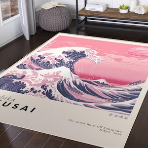 May include: A rectangular rug featuring a pink and navy blue depiction of "The Great Wave off Kanagawa" by Hokusai. The artwork includes text in English and Japanese, with the artist's name and the wave's title. The rug has a cream-coloured border.