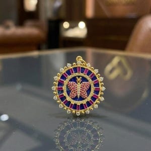 May include: A circular gold pendant featuring a central butterfly motif encrusted with pink stones. The butterfly is set against a deep blue background, surrounded by a ring of rectangular red stones and an outer border of small white stones.