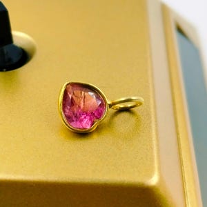 May include: A heart-shaped pendant with a pink and orange gemstone set in a gold-colored metal. The pendant has a small loop for a chain. The background is a gold-colored surface.